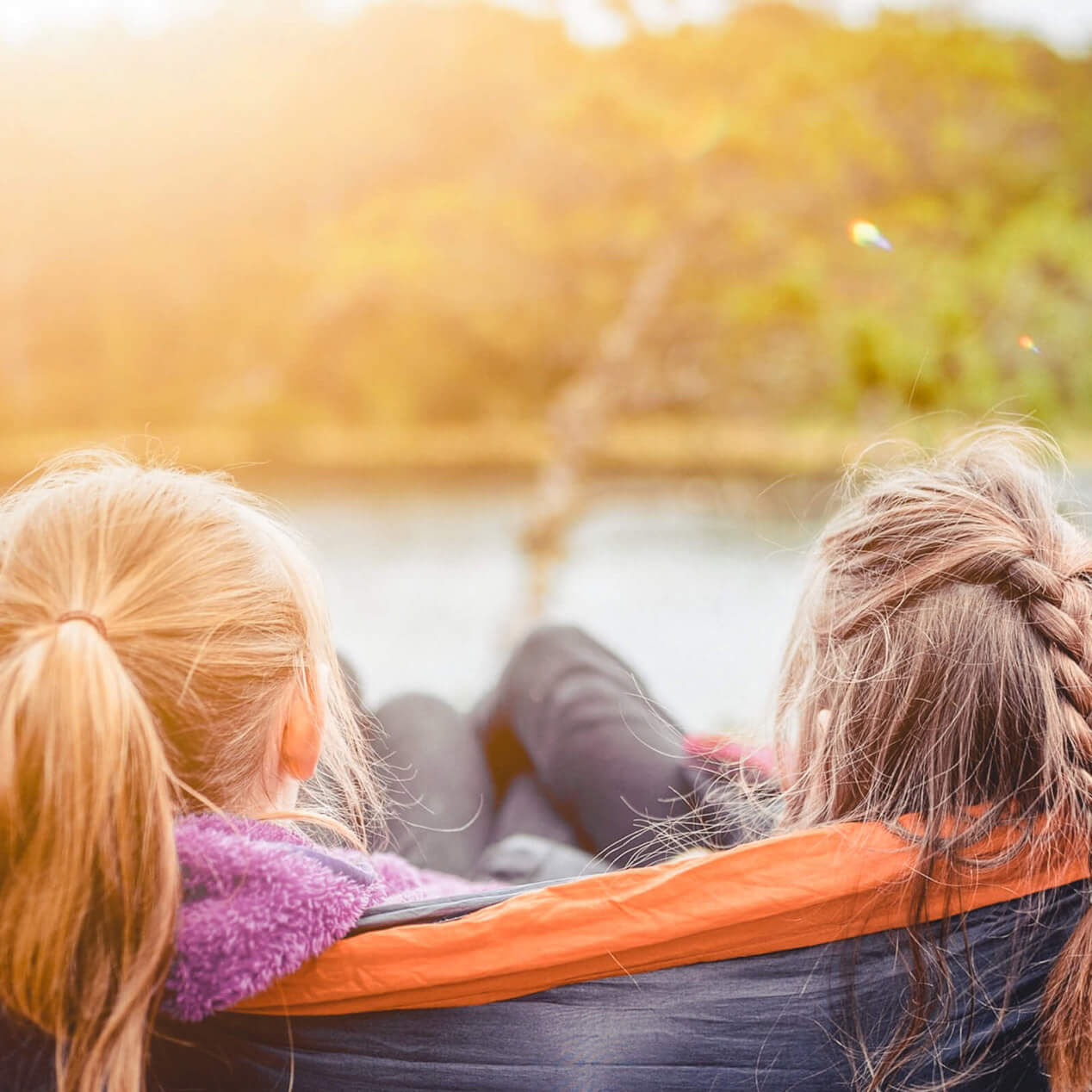 Zwei Mädchen liegen mit dem Hinterkopf zur Kamera in einer Hängematte. Sie schauen direkt auf einen See und die Sonne scheint.